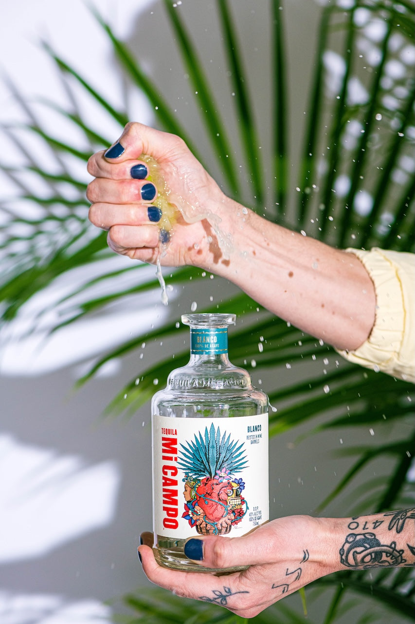 Mi CAMPO tequila bottle held with lime juice dripping, palm leaves in background.